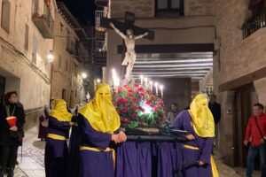 A group of people wearing purple robes and yellow hoods, carrying a statue of Jesus Christ on the cross while onlookers watch in silence during Semana Santa in Valderrobres.
